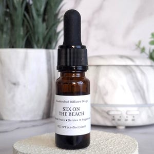 May include: A small amber glass bottle with a black dropper top. The label reads "Sex on the Beach" with "Natural Fruits • Berries • Sugar" below. The net weight is 0.34 fl oz (10ml). The bottle sits on a textured white surface.