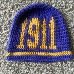 Puede incluir: Un gorro de ganchillo azul hecho a mano con el número "1911" en hilo dorado. El gorro tiene una franja dorada en la base. El artículo se muestra sobre una superficie gris texturizada. Es un accesorio hecho a mano.
