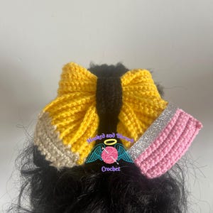 May include: A handcrafted crochet hair bow resembling a pencil. It features yellow, black, and beige yarn, with a pink eraser and silver glitter detail. The text "Hooked and Blessed Crochet" is visible.