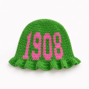 May include: A green crochet hat with the number "1908" in pink. The hat has a ruffled brim and is made of a textured yarn. The hat is displayed on a white background.
