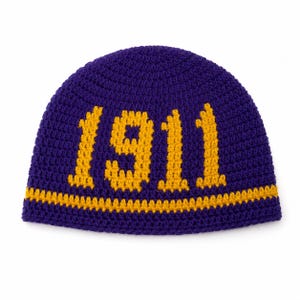 May include: A crocheted beanie in a deep purple color with the year "1911" in gold lettering. The hat features a gold stripe at the base. The beanie is likely handmade and suitable for cold weather.