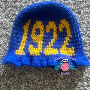 Könnte beinhalten: Blauer gehäkelter Fischerhut mit der gelben Zahl "1922". Der Hut hat einen gerüschten Rand und den Text "Hooked and Blessed Crochet" mit einer Garnkugel und Flügelgrafik.