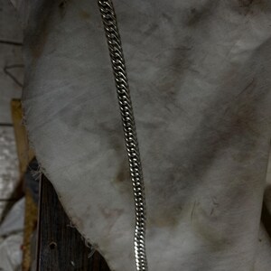 Puede incluir: Una pulsera de cadena de metal en tono plateado que presenta una serie de eslabones planos, pulidos y entrelazados dispuestos en un patrón uniforme y continuo. La pulsera es recta y muestra un brillo metálico en toda su longitud.