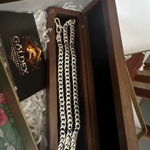 Puede incluir: Un collar de cadena de plata con diseño de eslabones de bordillo se muestra dentro de una caja de joyería de madera abierta. El interior de la caja es de terciopelo negro. También se ve una tarjeta de visita con el logotipo "Goldex Jewellery".