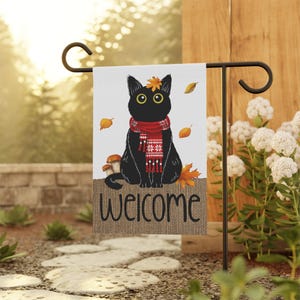 May include: A rectangular garden flag featuring a black cat wearing a red patterned scarf with a single orange leaf on its head. The flag displays the word "welcome" in black script over a burlap-textured base, surrounded by falling autumn leaves.