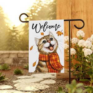 May include: A rectangular garden flag featuring a white background with the word Welcome in black script at the top. A brown tabby cat with a wide, happy expression wears a plaid orange and yellow scarf, surrounded by falling autumn leaves.