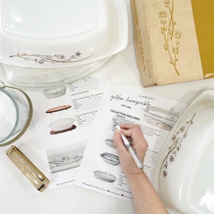 Può includere: Un elenco di controllo per una collezione vintage Pyrex Golden Honeysuckle. L'elenco di controllo include un elenco di articoli con caselle di controllo per spuntare ogni articolo. L'elenco di controllo è stampato su carta bianca con un motivo floreale dorato. L'elenco di controllo è su una superficie bianca con una casseruola Pyrex sullo sfondo.