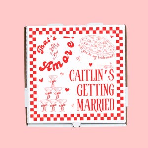 May include: A white pizza box with a red and white checkered pattern. The box has a red design with the text "That's Amore!" and a drawing of a cupid. The text "Will you be my bridesmaid?" is written next to a drawing of a pizza. The text "Caitlin's Getting Married" is also on the box.