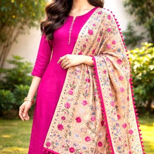 Könnte beinhalten: Ein magenta Salwar Kameez-Ensemble. Das Outfit umfasst eine lange Tunika mit Dreiviertelärmeln und passender Hose. Ein beiger Schal mit floraler Stickerei und rosa Bommelbesatz ist über den Schultern drapiert. Der Hintergrund ist eine verschwommene Szene im Freien.