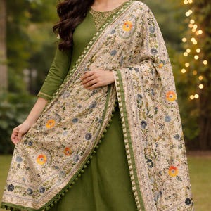Könnte beinhalten: Olivgrünes Salwar-Kameez-Ensemble mit einem floral bestickten Schal. Der Schal hat einen cremefarbenen Hintergrund mit bunten Blumenmustern und grünen Bommelbesatz. Das Outfit umfasst eine langärmlige Tunika und eine passende Hose.