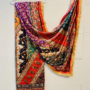 May include: A vibrant, patterned scarf featuring a mix of designs and colours, including red, purple, and gold. The scarf is suspended from a white hanger. The edges are trimmed with gold fringe.