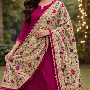Könnte beinhalten: Ein magenta Salwar Kameez-Ensemble. Das Outfit umfasst eine langärmlige Tunika und eine passende Hose. Ein beiger Schal mit Blumenstickerei in Rosa, Lila und Blau und mit magentafarbenen Bommeln verziert, ist über den Schultern drapiert.