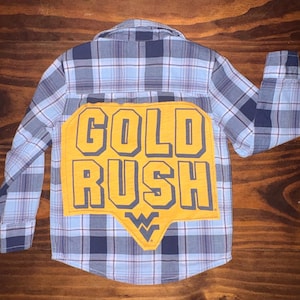 May include: A blue and white plaid button-down shirt with a large yellow patch on the back. The patch features the words "GOLD RUSH" in bold, dark blue letters, with the WVU logo at the bottom.