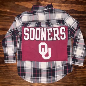 May include: A plaid button-down shirt in black, white, and pink, with a maroon patch featuring the word "SOONERS" in white block letters and a white "OU" logo. The shirt is laid flat.