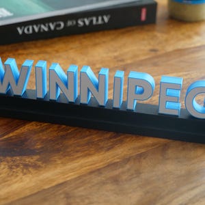 May include: A 3D sign reading "WINNIPEG". The letters are light blue with silver edging, mounted on a black base. The sign is displayed on a wooden surface.