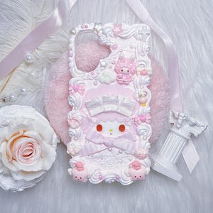 Peut inclure: Une coque de téléphone décorée d'une texture ressemblant à de la crème fouettée blanche, de rubans roses, de perles et de petites figurines, dont un lapin rose et un chat blanc. Le design présente un nœud rose central, de la dentelle et des étoiles.