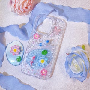 May include: Clear phone case with a pond design featuring pink and blue koi fish, lotus flowers, and lily pads. The case is decorated with pink flowers and glitter. A matching phone stand is also present.
