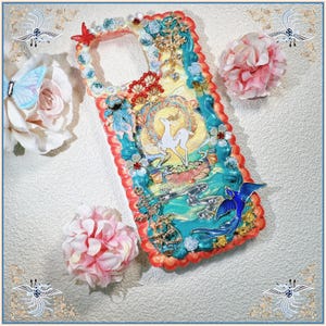 Peut inclure: Coque de téléphone ornée avec un design vibrant. Elle présente un cerf dans un emblème circulaire, entouré de motifs floraux et de vagues. Le boîtier a une bordure rouge, turquoise et dorée, avec un charm oiseau bleu et des embellissements décoratifs.