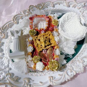 Puede incluir: Funda de teléfono ornamentada con un diseño rojo y dorado, adornada con rosas doradas, perlas y una mariposa. La funda presenta un relieve dorado decorativo y está colocada sobre una bandeja blanca ornamentada con un espejo.