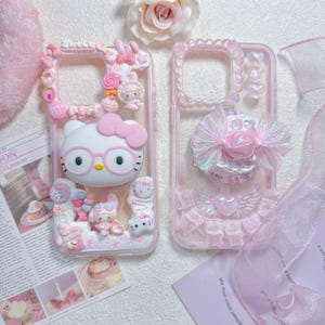 Puede incluir: Dos fundas de teléfono transparentes decoradas con adornos rosas y blancos. La funda izquierda presenta un personaje de gato blanco con gafas rosas, lazos y dijes. La derecha incluye un lazo iridiscente, un corazón y un borde de encaje.