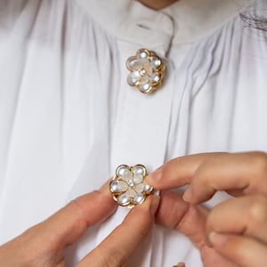 Peut inclure: Gros plan sur une chemise blanche avec des boutons en forme de fleur. Les boutons ont un bord doré, des pétales blancs et un petit bijou transparent au centre. La chemise est boutonnée.