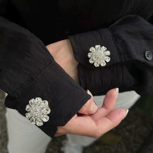 May include: A black shirt with three decorative crystal buttons. The buttons are in the shape of a flower and have a gold base.