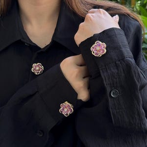 May include: Black button-down shirt with flower-shaped cuff links. The cuff links have a gold-coloured base with pink and clear gemstone accents. The shirt is made of a textured fabric.
