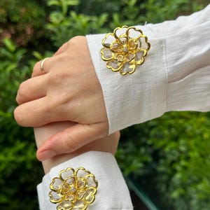 May include: Gold-toned flower-shaped cufflink set. Each cufflink features a clear, faceted center stone and a delicate, openwork design. The cufflinks are displayed on a white shirt cuff, showcasing their elegant details and craftsmanship.
