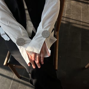 May include: A white long-sleeved shirt with large, round, rhinestone-studded cuff links. The shirt is paired with a black vest and pants. The person is seated in a wooden chair.