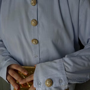 May include: A light blue button-down shirt with gold buttons featuring a pearl center. The shirt is worn by a person holding a gold clutch.