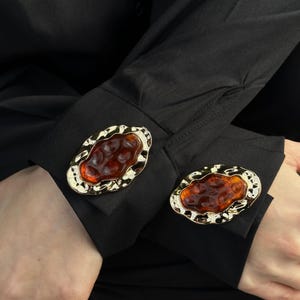 May include: A pair of gold-toned cufflinks with an oval, amber-colored center. The cufflinks are set against the black cuff of a long-sleeved shirt. The design features a textured gold border.