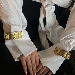 May include: Two gold cufflinks with a textured, swirling design. The cufflinks are on a white shirt with the sleeves rolled up.