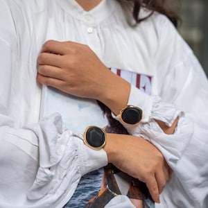 May include: Gold-toned cuff bracelets with large, black, oval-shaped stones. The bracelets are worn on the wrists of a person wearing a white blouse. The cuffs have a modern, minimalist design, suitable for everyday wear or special occasions.