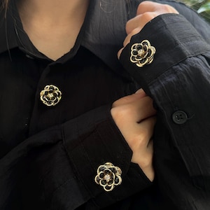 Può includere: Camicia nera con gemelli a forma di fiore dorati e neri. I gemelli hanno un bordo dorato, petali neri e una piccola gemma trasparente al centro. La camicia ha un colletto classico e polsini abbottonati.