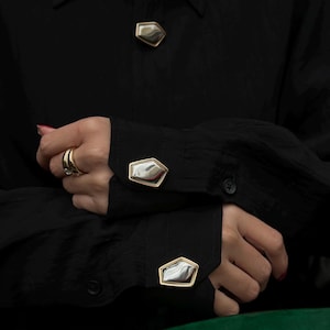 May include: Black button-down shirt with geometric silver and gold cuff links and a matching button. The cuff links and button have a unique, irregular shape. The shirt is made of a textured fabric.
