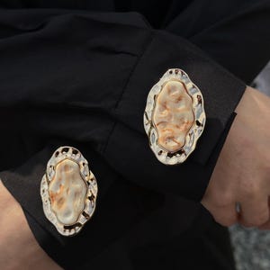 May include: Close-up of a pair of gold-toned cufflinks with an abstract, organic design. The cufflinks feature a textured, off-white center with brown and gold accents, set within a silver-colored frame. The cufflinks are on a black shirt.