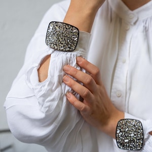 Op de afbeelding: Twee vierkante manchetarmbanden met een zwarte rand en een mozaïek van kleine, glanzende, zilverkleurige kristallen. De armbanden worden gedragen op de manchetten van een wit overhemd met lange mouwen.