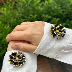 May include: Black and gold flower-shaped cufflinks with a small, clear gem in the center. The cufflinks are worn on a white shirt sleeve. The background is blurred green foliage.