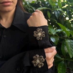 May include: Black long-sleeve shirt with flower-shaped cuff links. The cuff links are made of small, sparkling, dark-colored stones arranged in a floral pattern. The shirt has a classic button-down design.