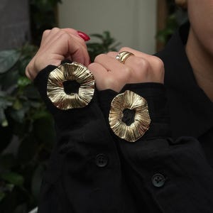May include: Gold-colored, textured cuff links in a floral design are attached to the sleeves of a black shirt. A gold ring with a clear stone is on a finger. The nails are painted red.