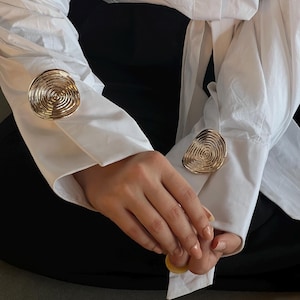 Peut inclure: Une chemise blanche boutonnée avec deux boutons décoratifs circulaires dorés sur les poignets.