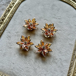 May include: Four flower-shaped earrings with orange and silver accents. Each earring features layered petals and a central crystal. The earrings are displayed on a light gray surface, framed by a gold-colored ornate border.