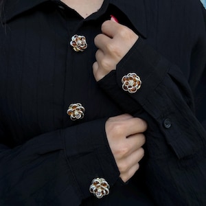 May include: Black button-down shirt with flower-shaped buttons. The buttons have a silver-colored base with amber-colored petals and a central crystal. The shirt's fabric has a subtle texture.