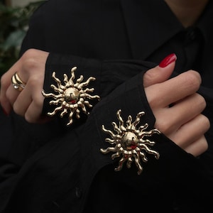 May include: Close-up of gold sunburst cufflinks on a black shirt. The cufflinks have a central sphere with radiating rays. The image focuses on the cuff detail, showcasing the jewellery's design against the dark fabric of the long-sleeved shirt.