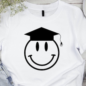 May include: White t-shirt featuring a black smiley face wearing a graduation cap. The smiley face has oval eyes and a curved smile. The graduation cap has a tassel hanging from it. The shirt is made of a soft material.