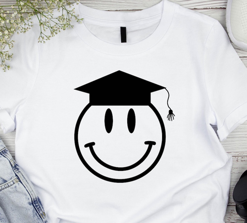 May include: White t-shirt featuring a black smiley face wearing a graduation cap. The smiley face has oval eyes and a curved smile. The graduation cap has a tassel hanging from it. The shirt is made of a soft material.