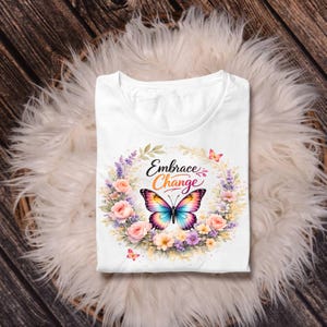 Puede incluir: Camiseta blanca con un dise&ntilde;o de mariposa colorida y las palabras "Embrace Change" en una corona floral. La mariposa tiene alas de arco&iacute;ris. La camiseta est&aacute; doblada sobre una superficie blanca y esponjosa.