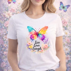 Puede incluir: Camiseta blanca con un dise&ntilde;o de mariposa en acuarela vibrante en tonos azules, amarillos, naranjas y morados. La mariposa est&aacute; rodeada de una corona floral y las palabras "Live Love Laugh".