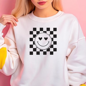May include: White sweatshirt featuring a black and white checkered pattern with a smiley face design. The smiley face has heart-shaped eyes and a curved mouth. The sweatshirt has a crew neck and long sleeves with a yellow accent on the cuff.