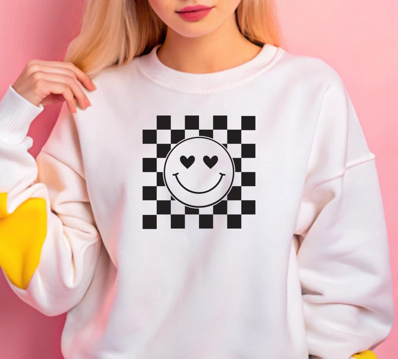 May include: White sweatshirt featuring a black and white checkered pattern with a smiley face design. The smiley face has heart-shaped eyes and a curved mouth. The sweatshirt has a crew neck and long sleeves with a yellow accent on the cuff.